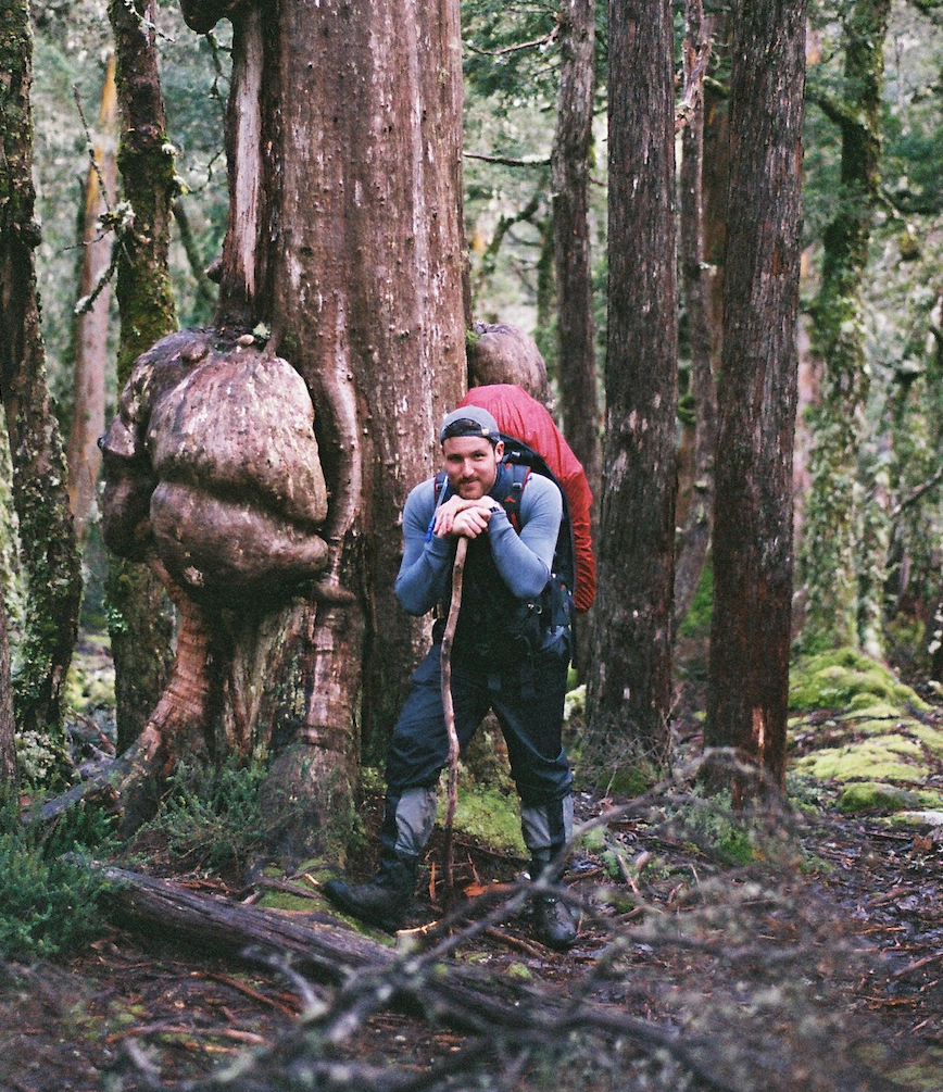 A photo of Brady with a hiking back pack, taking a short rest standing in front of a tree.